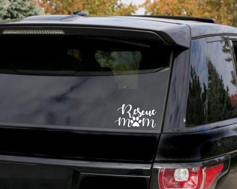 May include: White vinyl decal with the words "Rescue Mom" in a cursive font, with a paw print replacing the "o" in "mom". The decal is affixed to the rear window of a black SUV.