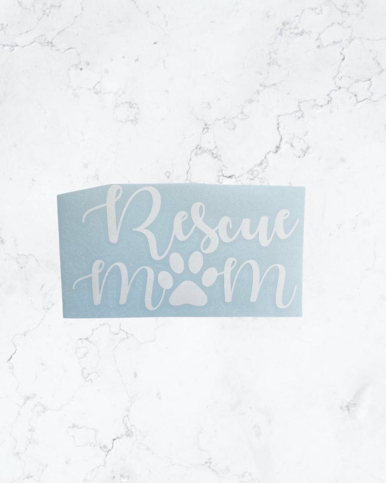 May include: A light blue sticker with the words "Rescue Mom" in white cursive script. A white paw print graphic is between the words "Rescue" and "Mom". The sticker is rectangular and sits on a white marble surface.