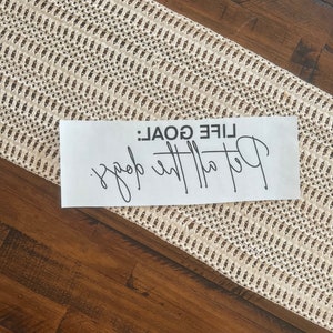 K&ouml;nnte beinhalten: Ein wei&szlig;es rechteckiges Schild mit dem Text "LIFE GOAL: Better the goal" in schwarzer Schrift. Das Schild liegt auf einem cremefarbenen Tischl&auml;ufer mit offener Webart auf einer dunklen Holzoberfl&auml;che.