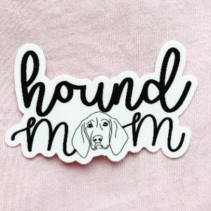 May include: White sticker with the words "hound mom" in a black, cursive font. A line drawing of a hound dog's face is centered below the word "hound". The sticker is on a pink background.