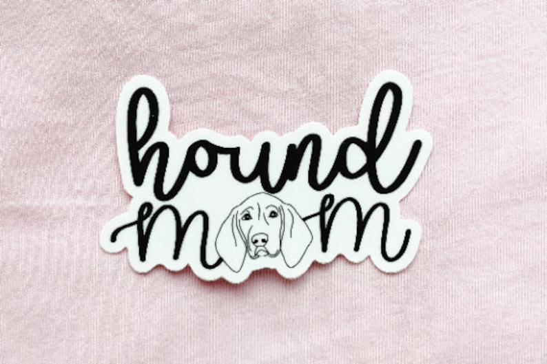 May include: White sticker with the words "hound mom" in a black, cursive font. A line drawing of a hound dog's face is centered below the word "hound". The sticker is on a pink background.