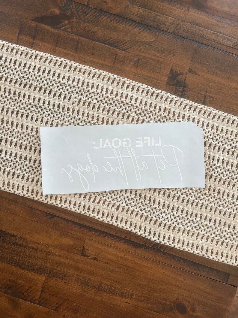 K&ouml;nnte beinhalten: Ein wei&szlig;es rechteckiges Abziehbild mit dem Text "Put the goal: LIFE GOAL" in Schreibschrift. Das Abziehbild befindet sich auf einer Holzoberfl&auml;che neben einem cremefarbenen Tischl&auml;ufer mit offener Webart.
