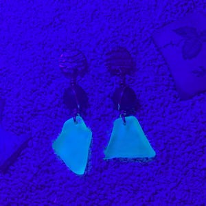 May include: A pair of drop earrings featuring dark, textured circular studs connected to dark spherical beads, which suspend translucent, irregular, light-blue glowing pieces of sea glass that fluoresce brightly under ultraviolet light.