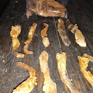 May include: A close-up of a wooden surface with several dried, brown, and orange pieces of food. The food is arranged in a row, and some of the pieces are in a plastic bag.