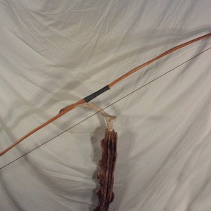 May include: A wooden bow with a black leather grip and a string attached. The bow is resting on a wooden stand.