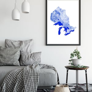 Peut inclure: Une impression encadr&eacute;e d'une silhouette bleue de la r&eacute;gion des Grands Lacs, sur fond blanc. L'impression est accroch&eacute;e &agrave; un mur blanc dans un salon, avec un canap&eacute; gris, une couverture et une table d'appoint avec une plante.