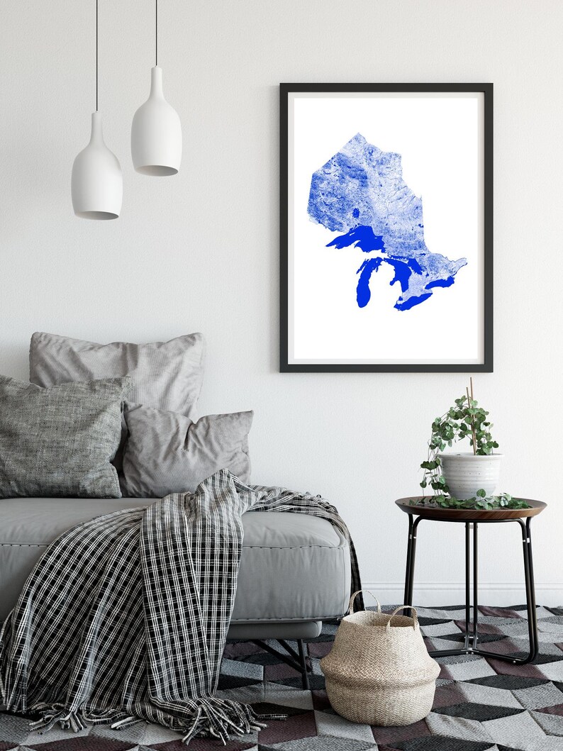 Peut inclure: Une impression encadr&eacute;e d'une silhouette bleue de la r&eacute;gion des Grands Lacs, sur fond blanc. L'impression est accroch&eacute;e &agrave; un mur blanc dans un salon, avec un canap&eacute; gris, une couverture et une table d'appoint avec une plante.