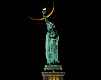 May include: The Statue of Liberty at night, illuminated against a black sky. The statue is a greenish-blue color, holding a torch with a bright flame. A crescent moon shape is visible above the statue.