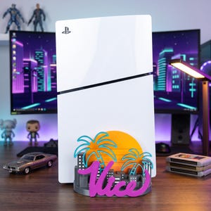 以下が含まれることがあります： 白いPlayStation 5用コンソールスタンド。装飾的なベースには、様式化されたオレンジ色の太陽、2本のティールカラーのヤシの木、灰色の都市のスカイライン、そして大きな紫色の筆記体で書かれた「Vice」という文字がデザインされています。
