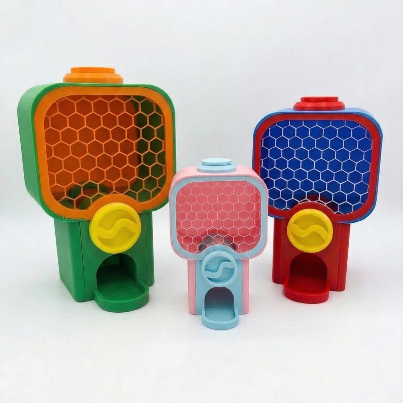 Puede incluir: Tres m&aacute;quinas de chicles coloridas en verde, rosa y rojo. Cada m&aacute;quina tiene una cara cuadrada con un patr&oacute;n de panal y una ranura para monedas amarilla. La m&aacute;quina verde tiene una cara naranja, la rosa tiene una cara rosa y la roja tiene una cara azul.