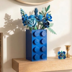 May include: A bright blue vase, designed to look like a building block, contains an arrangement of blue flowers. The vase is rectangular with eight circular studs on the front. A small blue flower decoration and a gold candle holder are on the wooden shelf.