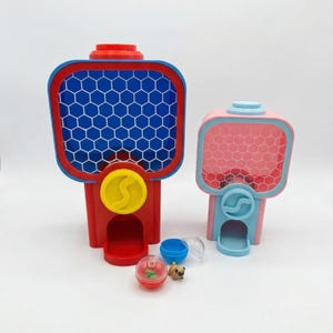 Puede incluir: Dos m&aacute;quinas de chicles de juguete de colores. Una es roja, azul y amarilla, y la otra rosa y azul. Las m&aacute;quinas tienen un patr&oacute;n de panal en la parte delantera. Peque&ntilde;os juguetes y recipientes est&aacute;n frente a las m&aacute;quinas.