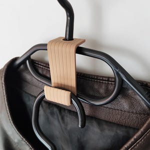 May include: A tan hanger connector is attached to a black plastic clothes hanger, holding a brown leather jacket. The connector has a ribbed design and is used to connect multiple hangers together.