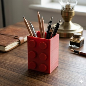 Puede incluir: Un organizador de escritorio rojo con forma de ladrillo, que contiene lápices y bolígrafos. El organizador tiene seis pernos circulares en relieve en la parte delantera. Los lápices son de madera con puntas de grafito y los bolígrafos son negros y plateados.