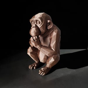 Könnte beinhalten: Eine bronzefarbene, geometrische Skulptur eines Schimpansen in sitzender Position, die Hände vor dem Gesicht gefaltet. Die Skulptur hat ein facettiertes Design und ist vor einem dunklen Hintergrund platziert.