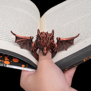 May include: A bronze-colored dragon book page holder. The dragon has detailed wings and a textured body, and is holding a book open. The dragon is a decorative item for book lovers.