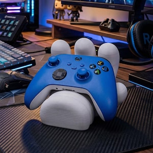 May include: A blue Xbox controller sits on a white hand-shaped stand. The controller features a black directional pad and coloured buttons. The stand is on a black surface, with gaming accessories in the background.