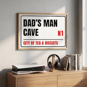 May include: A framed sign with the text "DAD'S MAN CAVE CITY OF TEA & BISCUITS N1" in black and red. The sign is mounted on a light-colored wall above a wooden cabinet. A gaming console, headphones, and books are on the cabinet.