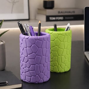 Peut inclure: Deux porte-stylos cylindriques, l'un violet, l'autre vert vif. Ils présentent tous deux un motif texturé en forme de mosaïque. Des stylos et des crayons sont visibles à l'intérieur. Les porte-stylos sont posés sur une surface sombre, comme un bureau.