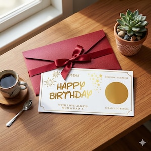 以下が含まれることがあります： 「Happy Birthday」の金色の文字とスクラッチオフエリアが付いた誕生日カード。赤いリボンが付いた赤い封筒、コーヒーカップ、小さな鉢植えの多肉植物の隣の木製のテーブルに置かれています。