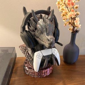 Puede incluir: Un soporte para auriculares con forma de dragón en tonos grises y negros, que sostiene unos auriculares negros y un mando de juego blanco. El soporte tiene un diseño detallado y texturizado y está colocado sobre una superficie de madera.