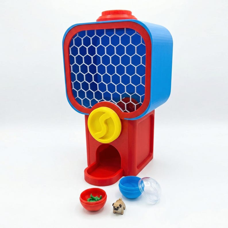 Puede incluir: Una m&aacute;quina de chicles roja, azul y amarilla con una ventana con patr&oacute;n de panal. La m&aacute;quina tiene una base roja, una parte superior azul y una ranura para monedas amarilla. Se muestran peque&ntilde;os juguetes y c&aacute;psulas debajo.