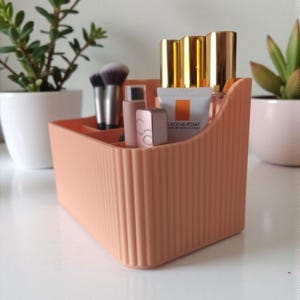 May include: A peach-colored, ribbed plastic organizer filled with makeup brushes, lipsticks, and skincare products. The organizer has multiple compartments and a curved front. Gold-capped bottles and a tube of "ROCHE-POSAY" are visible.