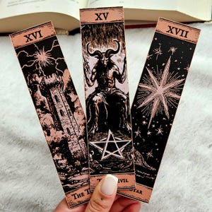 May include: Three tarot card bookmarks with black and rose gold illustrations. The cards feature images of a tower, a horned figure, and a star. The top of each bookmark has a Roman numeral.