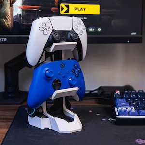 Könnte beinhalten: Ein weiß-schwarzer Gaming-Controller liegt auf einem blauen Gaming-Controller, beide auf einem weiß-schwarzen Ständer. Der Ständer steht auf einer schwarzen Oberfläche, mit Tastatur und Monitor im Hintergrund.
