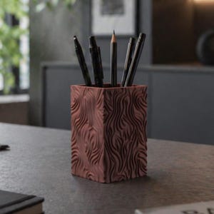 May include: A square, reddish-brown pen holder with a textured, wave-like pattern. The holder contains several black pens and a pencil. The pen holder sits on a dark gray surface, suggesting an office or workspace setting.