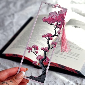 以下が含まれることがあります： 桜の木のデザインが施された、ピンク色のフレームの長方形のブックマーク。木にはピンクの花と黒い幹があります。ピンクのタッセルが上部にぶら下がっています。ブックマークは開いた本の中にあります。