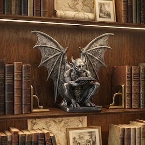 Puede incluir: Una detallada estatua de gárgola plateada con grandes alas texturizadas, sentada y leyendo un libro. La estatua se exhibe en una estantería de madera, rodeada de libros antiguos y obras de arte enmarcadas.