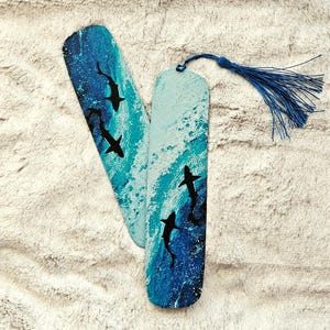 Op de afbeelding: Twee houten bladwijzers met een blauw oceaan golf ontwerp en zwarte haai silhouetten. Eén bladwijzer heeft een blauwe kwast bevestigd. De bladwijzers zijn beschilderd met tinten blauw en wit, waardoor een golfeffect ontstaat.