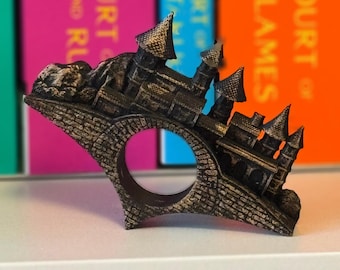 May include: A detailed, bronze-colored ring shaped like a castle. The ring features a castle design with multiple turrets, a bridge, and textured brickwork. The ring is set against a blurred background of colorful book spines.