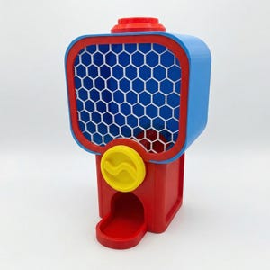 Puede incluir: Una m&aacute;quina de chicles roja, azul y amarilla. La parte superior es azul con un borde rojo y un patr&oacute;n de panal blanco. Un dial amarillo est&aacute; en la parte delantera. La base es roja.