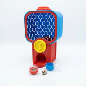 Puede incluir: Una m&aacute;quina de chicles roja, azul y amarilla con un dise&ntilde;o de panal en la parte delantera. Incluye una peque&ntilde;a figura de perro pug, una taza azul y una bola de pl&aacute;stico transparente con base roja.