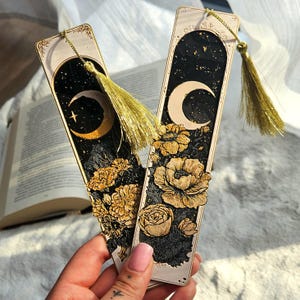 Puede incluir: Dos marcadores decorativos con borlas doradas. Cada marcador presenta un fondo negro con un diseño de luna creciente dorada y estrella. Un marcador tiene un diseño floral en la parte inferior. El otro marcador tiene un diseño floral en la parte inferior.