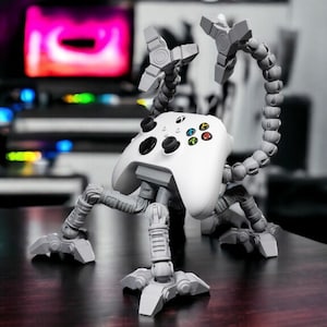 Könnte beinhalten: Ein weißer Xbox-Controller wird von einem grauen, beweglichen Ständer gehalten. Der Ständer hat mehrere Gelenke und einen flexiblen Hals mit einem Sockel für Stabilität. Der Controller hat schwarze Tasten und farbige Aktionstasten.