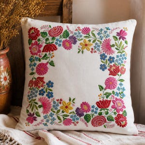May include: A square white throw pillow featuring a colorful embroidered floral border. The design consists of various flowers in shades of red, pink, yellow, purple, and blue, accented with green leaves and stems arranged in a rectangular frame.