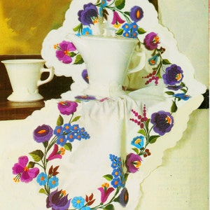 May include: A white tablecloth with a scalloped edge is decorated with embroidered flowers in shades of purple, blue, and pink. A white pitcher and a white cup are placed on the tablecloth.