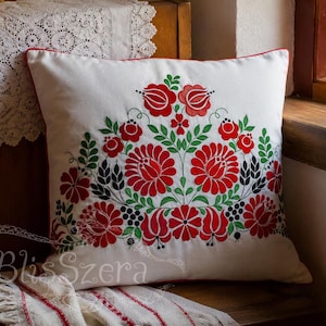May include: A square white throw pillow featuring a vibrant embroidered floral pattern in shades of red, green, and black. The design includes stylized flowers and leaves arranged in a symmetrical arc. The text BlisSzera is visible in the lower left.