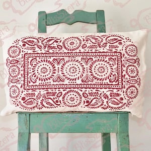 May include: A rectangular cushion with a white base and a burgundy floral design. The cushion is placed on a green wooden chair, highlighting the detailed pattern. The design features circular and paisley motifs.