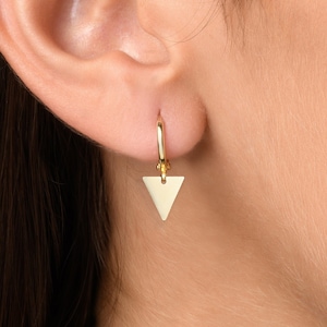 May include: A gold triangle earring with a hoop design. The earring is worn in the left ear of a person with long brown hair.