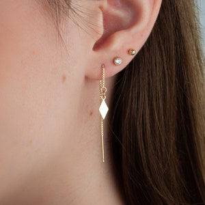 Puede incluir: Pendientes de cadena de oro con un dije en forma de diamante que cuelga de uno de los pendientes. Los pendientes se llevan en la oreja de una persona.