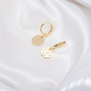 Peut inclure: Une paire de boucles d'oreilles créoles dorées avec des breloques en forme de disque martelé. Les boucles d'oreilles sont présentées sur un tissu blanc et soyeux. Elles ont un design simple et élégant.