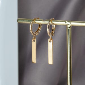 May include: Gold-coloured hoop earrings with rectangular bar pendants. The earrings are displayed on a gold-coloured jewellery stand, highlighting their design and metallic finish. A simple, elegant accessory.