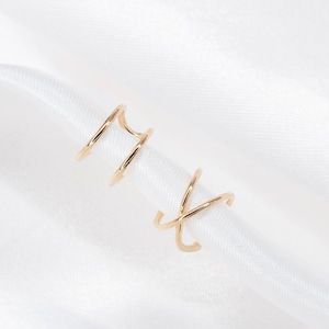 May include: Gold-colored ear cuffs with a spiral design. The ear cuffs are made of thin metal and have a minimalist style. The design features two separate pieces that wrap around the ear. The ear cuffs are displayed on a white fabric background.