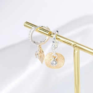 May include: A pair of silver hoop earrings with a dangling gold charm featuring a small clear stone. The earrings are displayed on a gold stand.