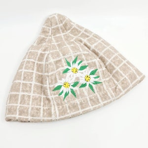 Könnte beinhalten: Ein beiger Häkelhut mit einem weißen Karomuster und gestickten weißen Edelweißblumen mit grünen Blättern.