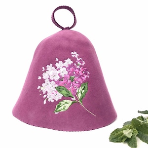 Könnte beinhalten: Eine mauvefarbene Sauna-Mütze mit einer floralen Stickerei aus weißen und lila Blüten und grünen Blättern. Die Mütze hat eine abgerundete Oberseite mit einer Schlaufe zum Aufhängen. Der Hintergrund ist weiß.